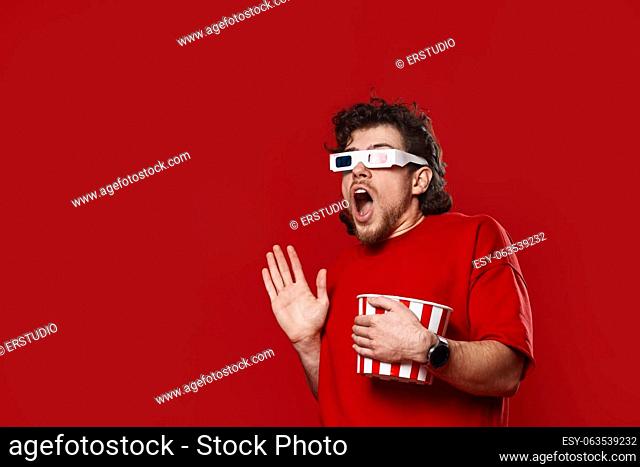 Shocked scared handsome man in 3d-glasses watching movie film hold bucket of popcorn on red background. copy space