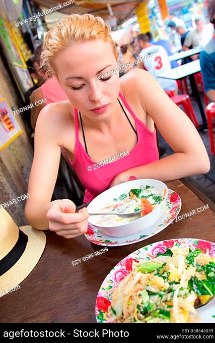 Young carefree adult female traveler eating traditional thai Tom Yum soup from the street stall vendor on Khao San road, Bangkok, Thailand