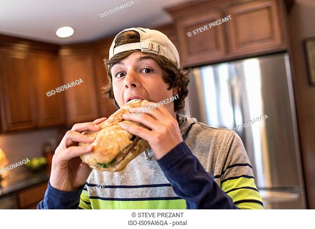 Teenage boy in kitchen biting large sandwich