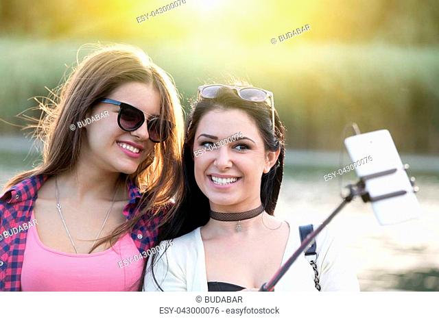 Two pretty teenage girls taking selfie on mobile phone with stick in the nature beside river. Positive emotions