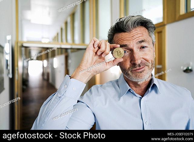 Businessman covering eyes with bitcoin while standing at office