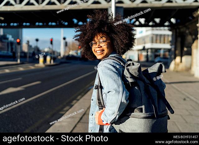 Smiling afro woman with backpack standing on sidewalk in city during sunny day