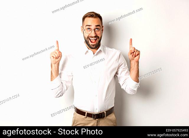 Excited businessman checking out advertisement, pointing and looking up with happy face, standing against white background