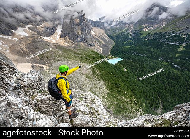 Young man points into the distance, mountaineer on a via ferrata Vandelli, view of Lago di Sorapis, Sorapiss circuit, mountains with low clouds, Dolomites