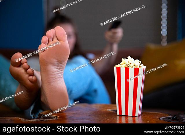 Beautiful young woman sitting on a living room couch, watching television with a red and white striped cardboard box with popcorn retro popcorn box movie night