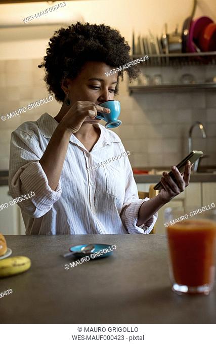 Portrait of young woman drinking espresso in her kitchen while looking at phablet