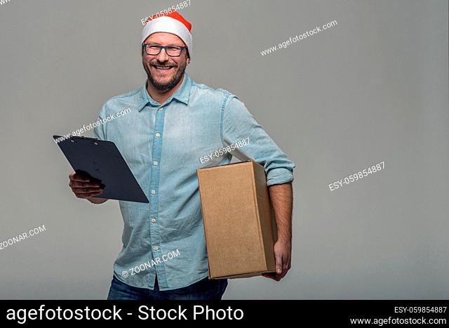Cheerful attractive young man in a Santa Hat carrying or delivering a large brown cardboard box for Christmas, over grey with copy space