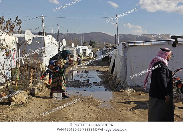 30 January 2019, Lebanon, Al Marj: A Syrian refugee woman carries her child while crossing a muddy path that runs along the tents of Al Marj refugee camp
