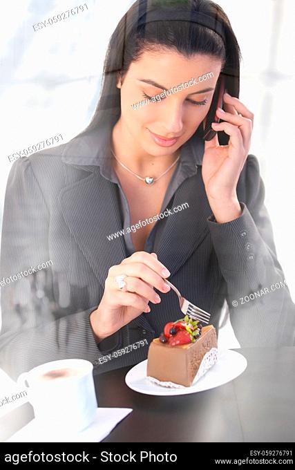 Smart office worker woman on phone call, sitting in cafe, having chocolate cake and coffee, smiling, picture through window
