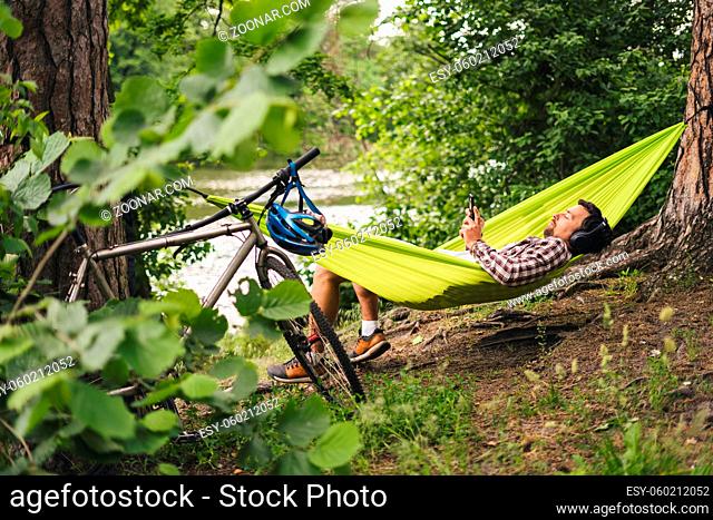 Man on bicycle trip at camping by lake is relaxing in green hammock while listening to music. Active recreation theme in nature