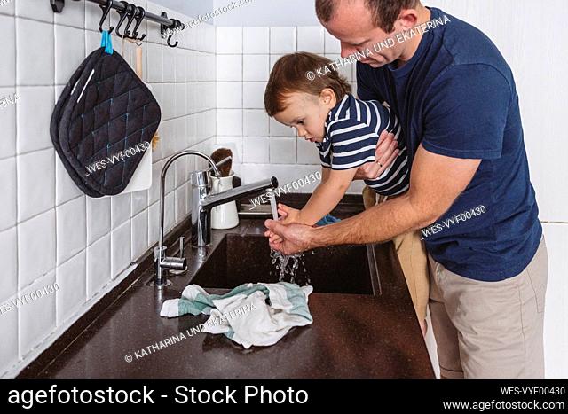Father carrying son while washing hands through faucet in kitchen at home
