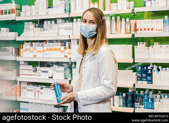Pharmacist wearing protective face mask while holding sanitizer against medicine shelf in chemist shop