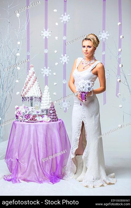Beautiful young woman in wool wedding dress holding bridal bouquet standing near small table with big wedding croquembouche cake