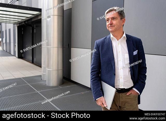 Contemplative mature businessman standing with laptop at footpath