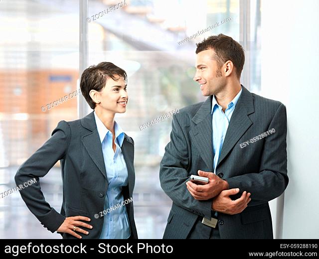 Two happy businesspeople talking in office lobby, looking at each other, smiling