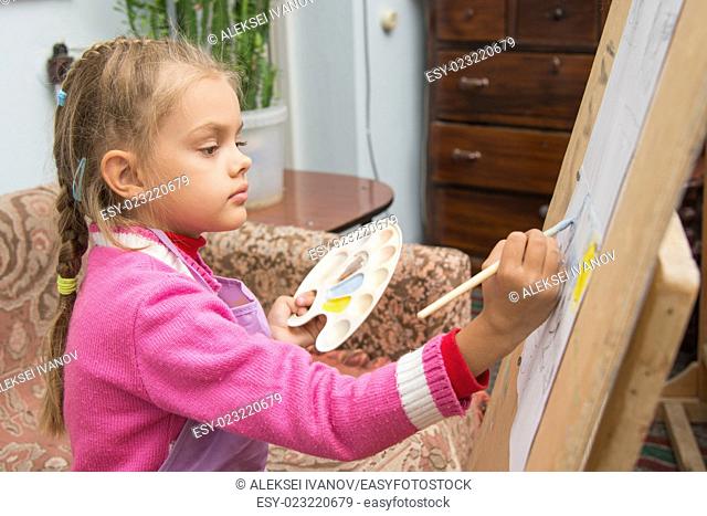 Five-year girl with a brush paints a picture on an easel and holding in his other hand in the artist's palette