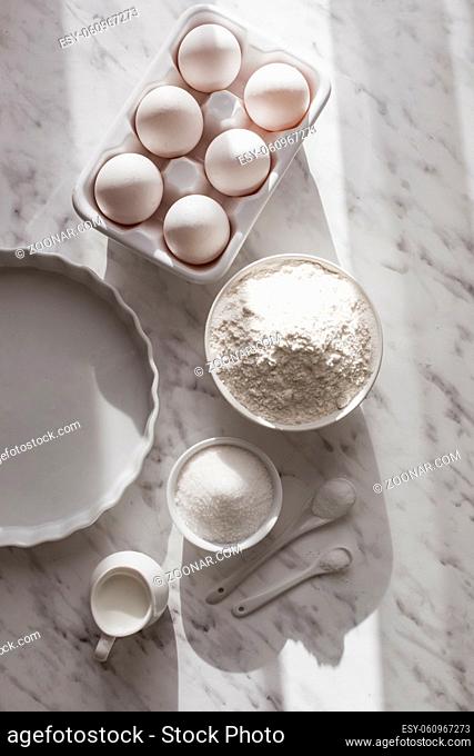 Top view ceramic tray with eggs placed near bowl of flour and sugar. White utensils with white ingredients on white surface. Copy space