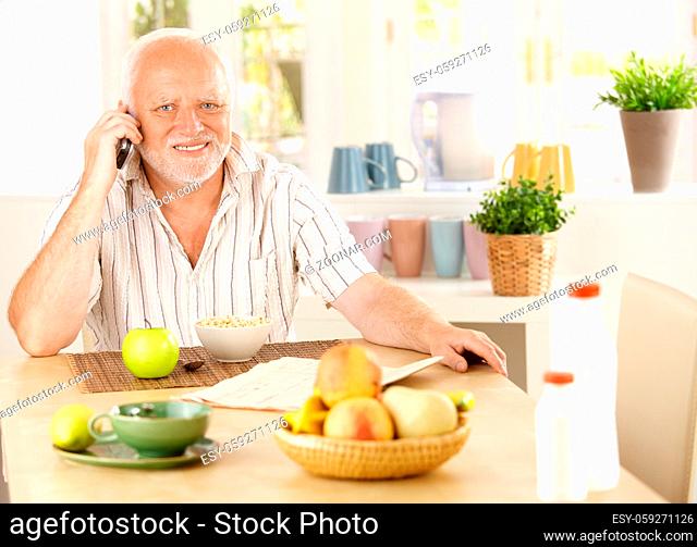 Healthy pensioner using cellphone at breakfast table, smiling at camera