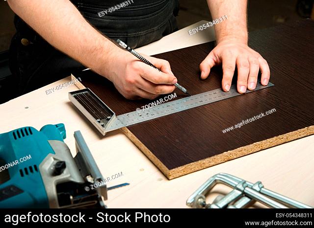 Men's hands draw a pencil on the ruler with a sub-standard template for cutting out furniture details next to the jigsaw and furniture clips