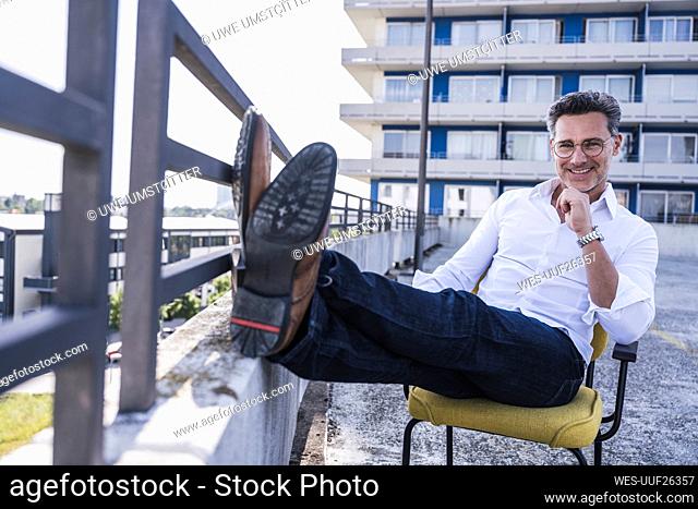 Smiling businessman with feet up sitting by railing on building terrace