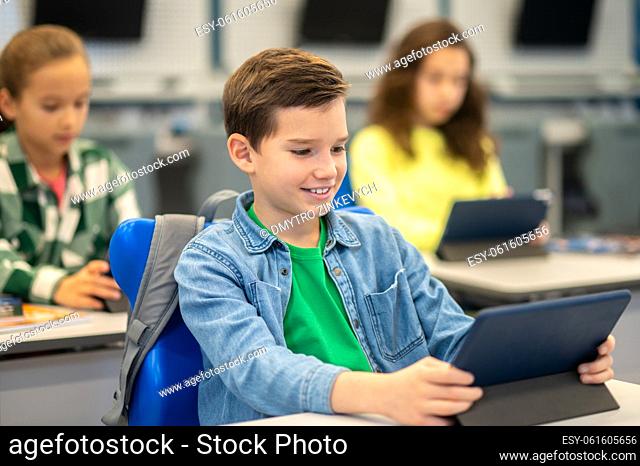 Studies. Smiling caucasian boy in casual clothes sitting at desk looking at tablet at school lesson and girls behind