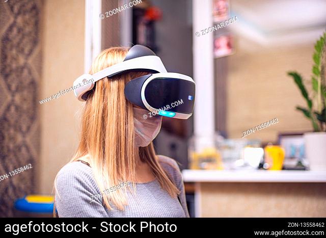 A young blond caucasian sick girl covers her face and wears a Virtual Reality Headset enjoys virtual reality in her apartment