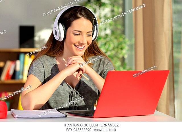 Portrait of a beautiful student viewing and listening video tutorials on line with headphones and a red pc sitting in a table at home