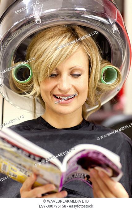 Happy woman reading a magazine with hair curlers under a hairdryer in a hairdressing salon
