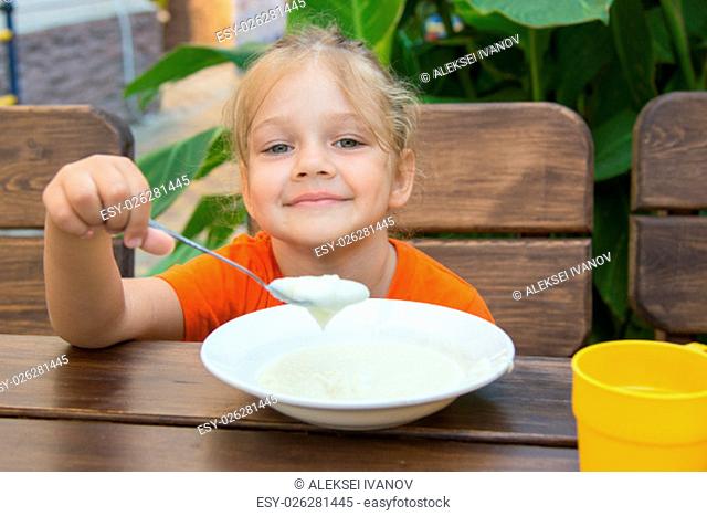 Five-year girl sits at a table on the veranda and enjoy eating porridge