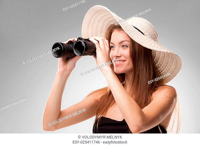Young woman in hat looking through binoculars isolated on gray background. Travel and adventure concept. Closeup