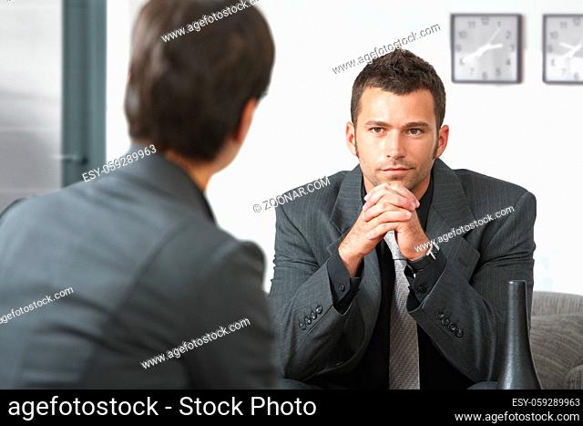 Young businessman sitting on sofa at office listening to talking businesswoman