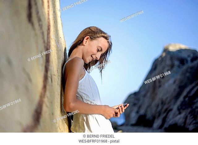 Croatia, Lokva Rogoznica, smiling girl leaning against rock looking at cell phone
