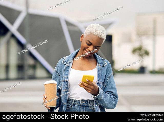 Smiling woman using smart phone holding disposable cup