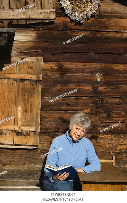 Austria, Senior woman reading book by log cabin