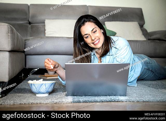 Smiling woman with laptop eating popcorn while lying on carpet at home
