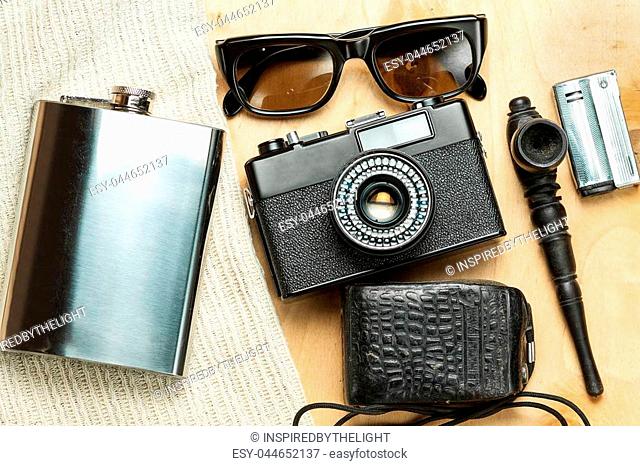Accessories of the creative person. 35-mm film camera, exposure meter in leather case, steel flask, sunglasses and smoking set on wooden background
