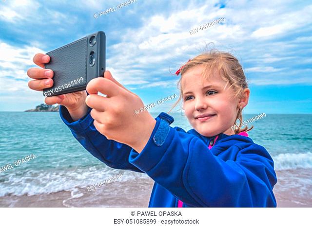 Cute little blond Caucasian girl taking selfie with mobile phone on the beach in autumn