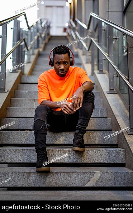 young african-american man in stylish orange hoodie sweatshirt in wireless headphones listening music and sitting on stairs