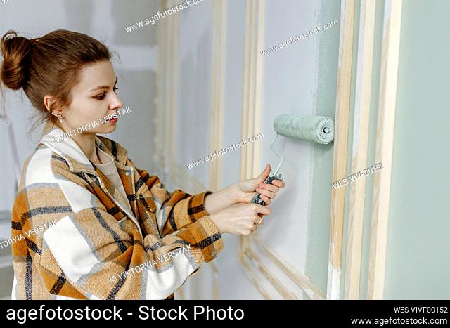 Young woman applying paint on wall renovating home