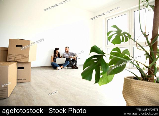 Man sitting with wife using laptop on hardwood floor in new unfurnished house