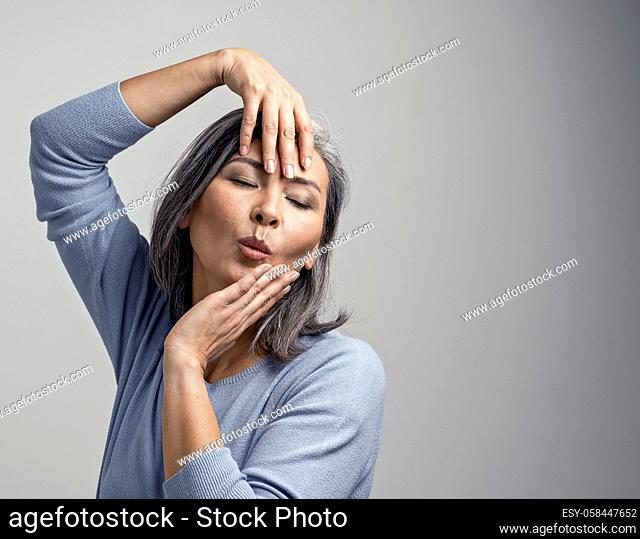 Beautiful Grey-Haired Female Model Posing For The Camera. Charming Middle-Aged Asian Woman Touching Her Face With Hands While Posing At The Photo Studio
