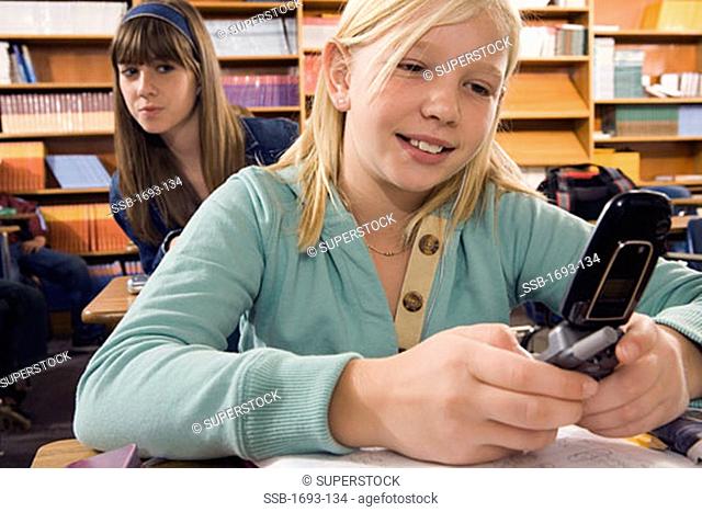 Girl using a mobile phone in a classroom with another girl looking over her shoulder