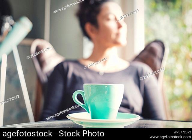 Hispanic woman at home sitting on modern chair in front of window relaxing in her living room and drinking tea, selective sharpness