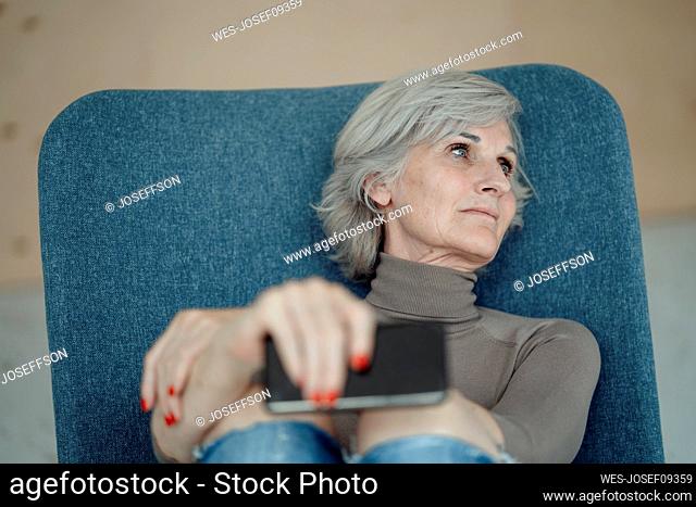 Thoughtful senior woman with smart phone sitting on chair in front of wall