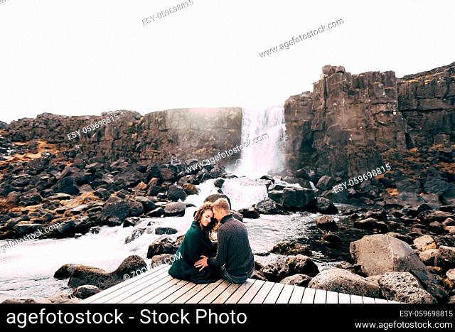A guy and a girl sit hugging on a bridge near Ehsaraurfoss Falls, Ehsarau River, National Park, Tingvedlir, Sudurland Region, Iceland