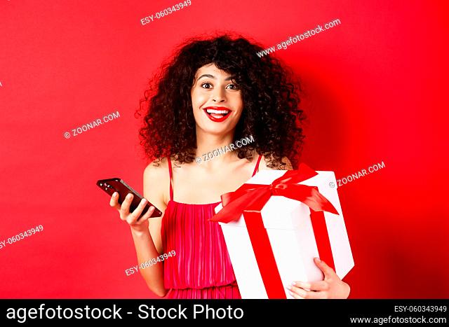 Online shopping and Valentines day. Beautiful young woman holding smartphone and lovers gift, looking surprised and happy at camera, red background