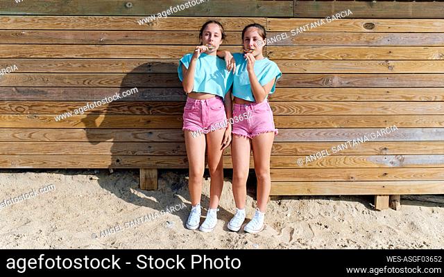 Twin sisters eating lollipops and standing in front of wooden wall