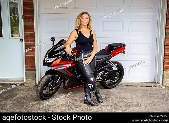 twenty something blond woman wearing leather pant and a black tank top, doing a duck face, while leaning on a sport motocycle