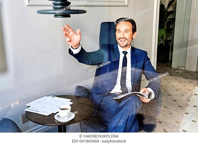 Hello. Happy man is waving arm to passerby through window while sitting in cozy cafeteria. He is holding business journal and laughing