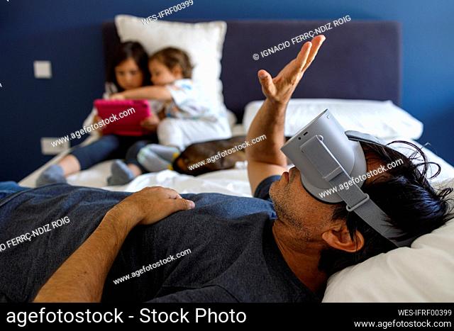 Man with virtual reality simulator gesturing while siblings using digital tablet on bed at home